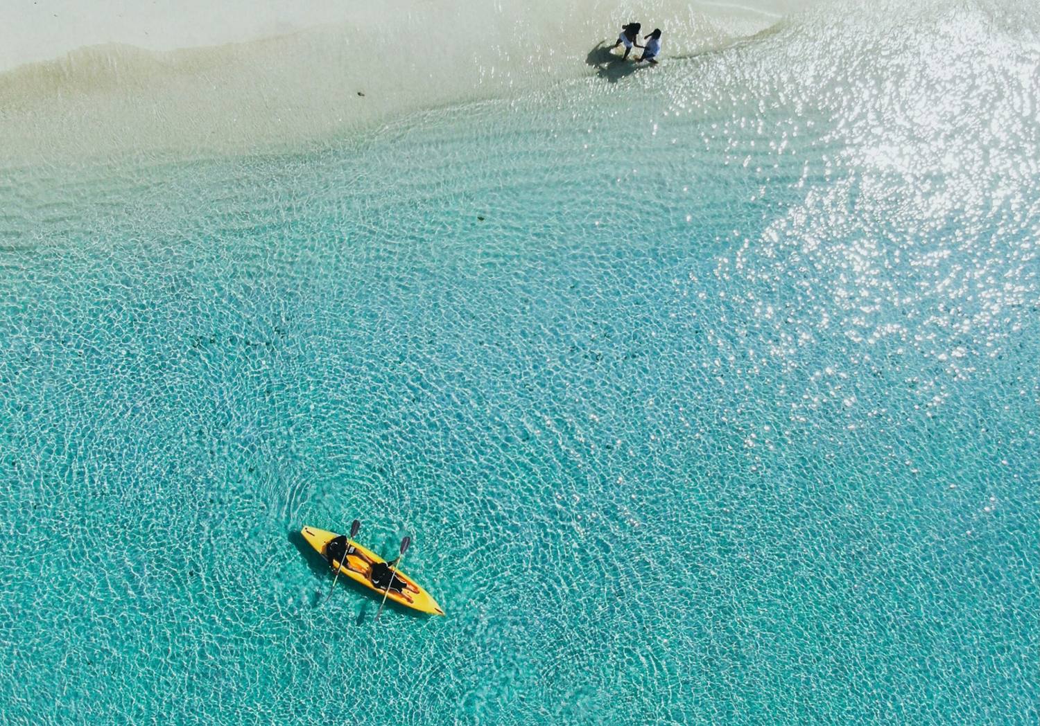 Aerial view of two people kayaking in clear turquoise water near a sandy shore